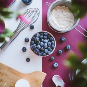 A-flat-lay-of-fresh-blueberries-in-a-bowl-a-sifter-with-flour-and-a-whisk-surrounded-by-baking-tools-and-ingredients.