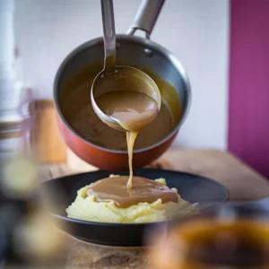 A-saucepan-of-homemade-gravy-being-whisked-with-a-ladle-pouring-the-gravy-over-a-plate-of-mashed-potatoes