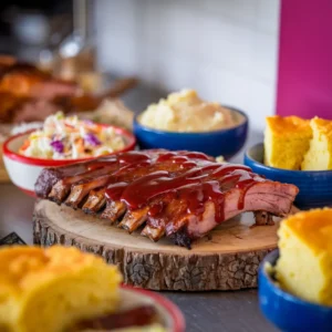 A-serving-of-ribs-on-a-rustic-wooden-board-surrounded-by-bowls-of-coleslaw-mashed-potatoes-and-cornbread-with-barbecue-sauce-drizzled-over-the-ribs